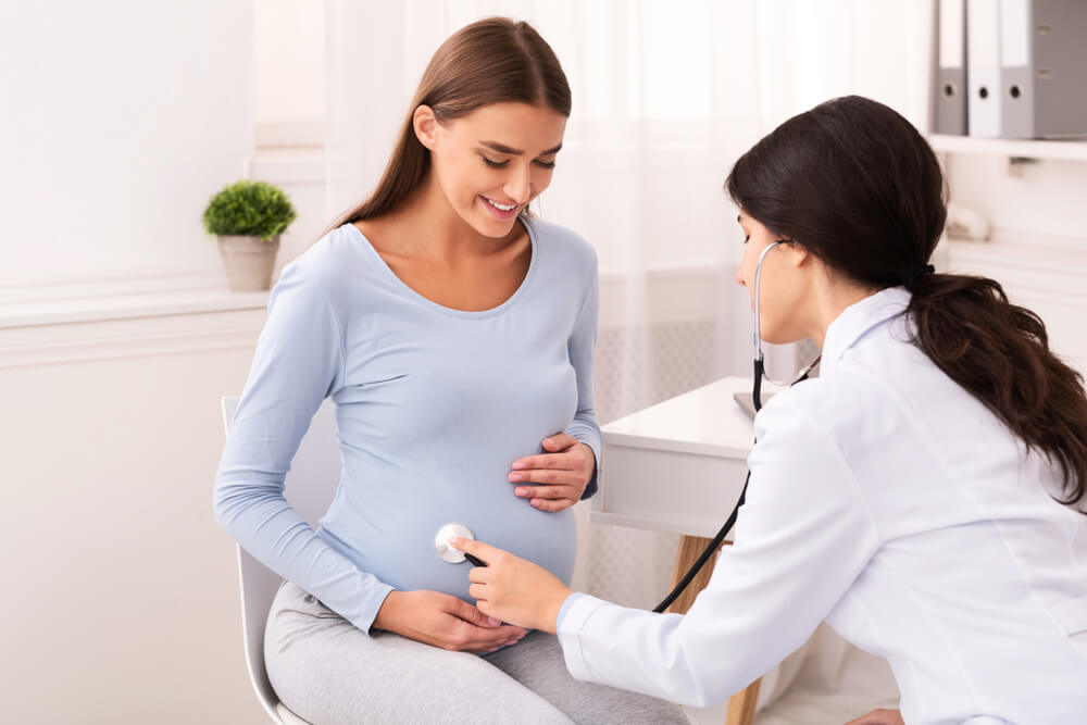 Doctor Examining Pregnant Woman Holding Stethoscope Near Belly Listening Baby’s Heartbeat in Office. Pregnancy Checkup