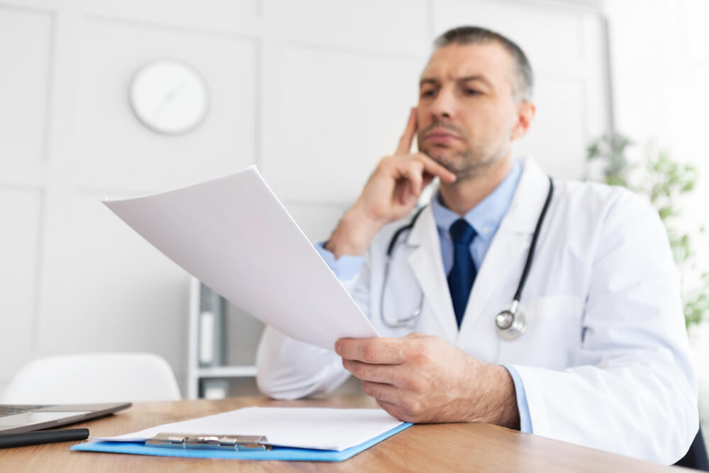 Male General Practitioner Holding Paper, Reading Medical Report, Sitting at Desk in His Office at Clinic