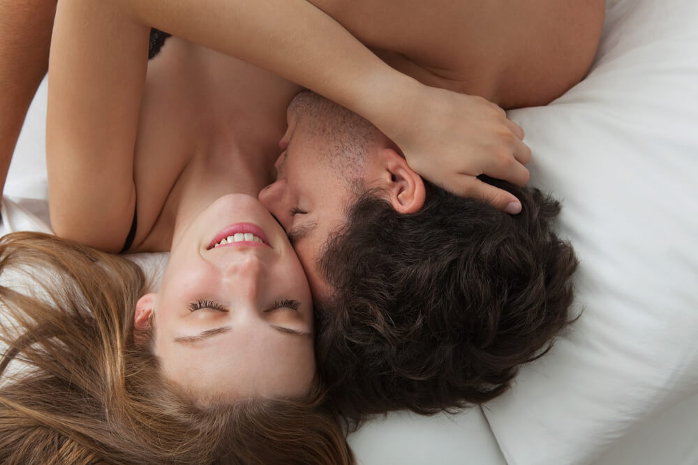 Overhead Close Up Portrait of a Young Romantic Couple Hugging and Kissing, Laying Down on a White Bed