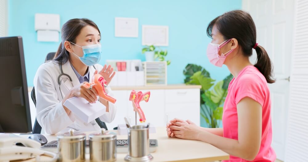 Asian Female Doctor Wear White Coat and Face Mask Explaining Uterus Model to Woman in Hospital