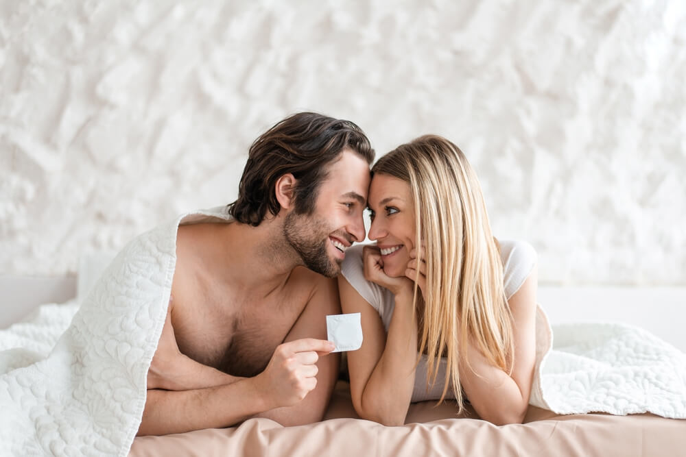 Young Spouses Holding Condom, Lying on Bed Under Blanket, Looking at Each Other With Love at Home