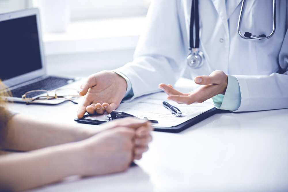 Doctor and Female Patient Sitting at the Desk and Talking in Clinic Near Window.