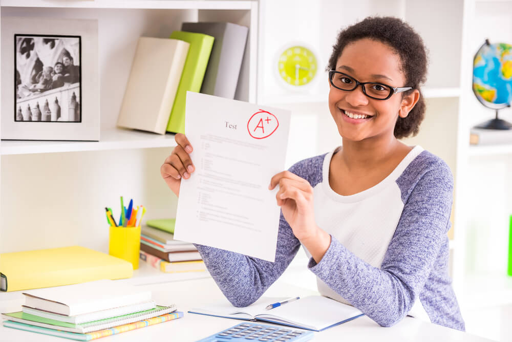 Young Schoolgirl in Glasses Sitting At the Table and Showing Perfect Test Results on Colorful Background.