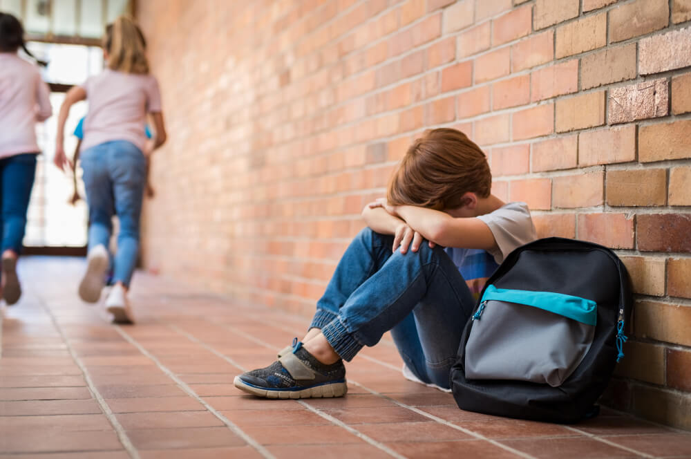 Little Boy Sitting Alone on Floor After Suffering an Act of Bullying While Children Run In the Background. 