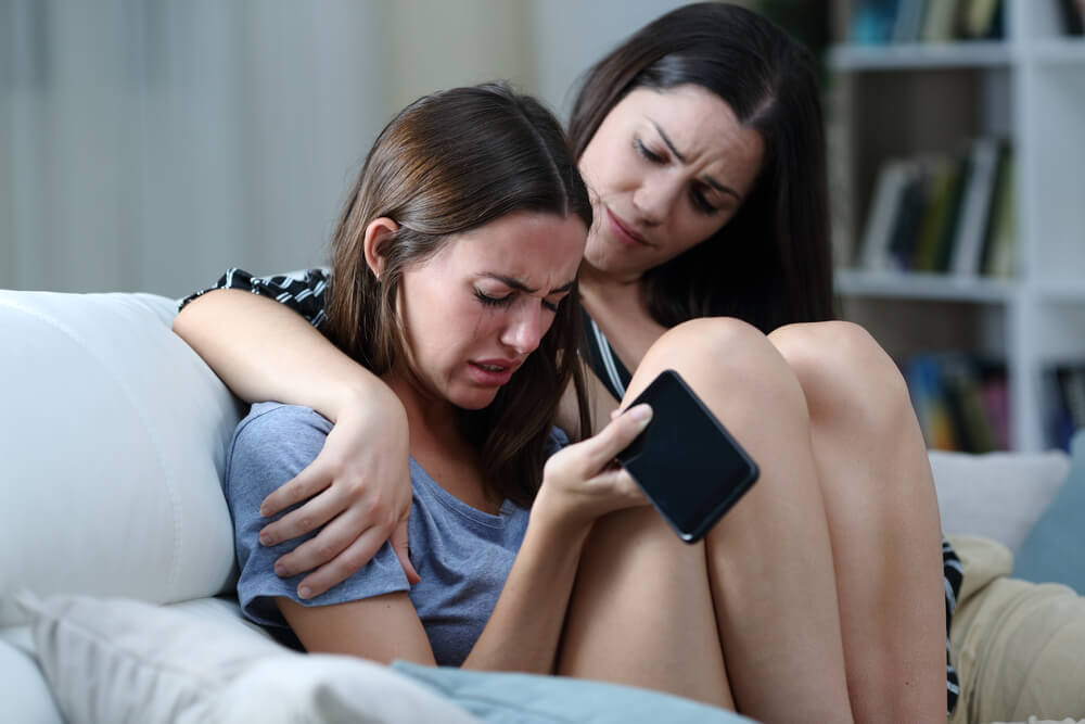 Sad Teen With Phone Being Comforted by Her Sister on a Couch in the Living Room at Home